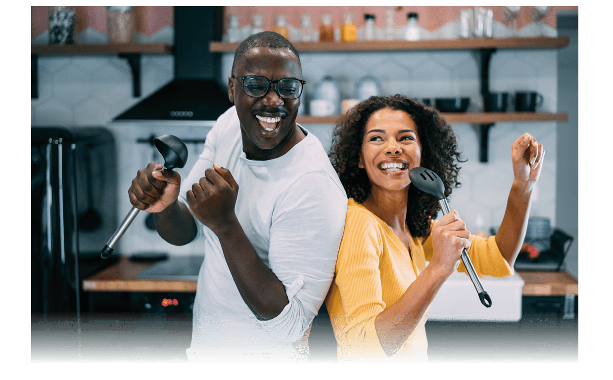Shot of happy couple having fun during cooking in the kitchen at home. Lovely african-american couple holding kitchen utensil and singing during a cooking.