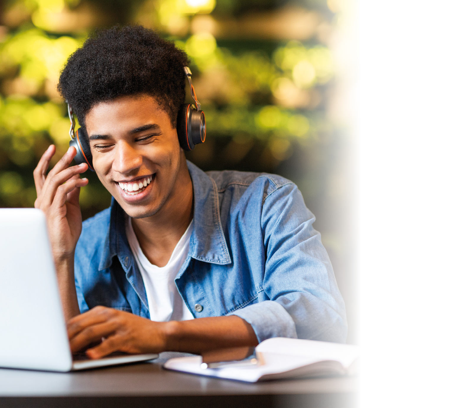 Cheerful black young guy with headset looking at laptop, having fun while studying, cafe interior