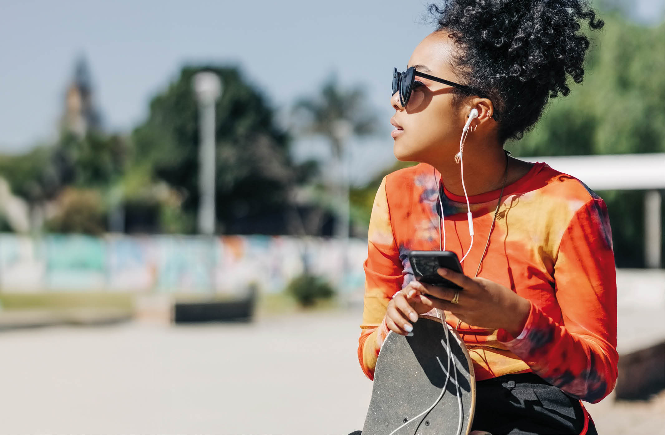 Young girl relaxing with her skateboard listening to music. Urban girl enjoying listening to music from her mobile phone while sitting at skatepark.