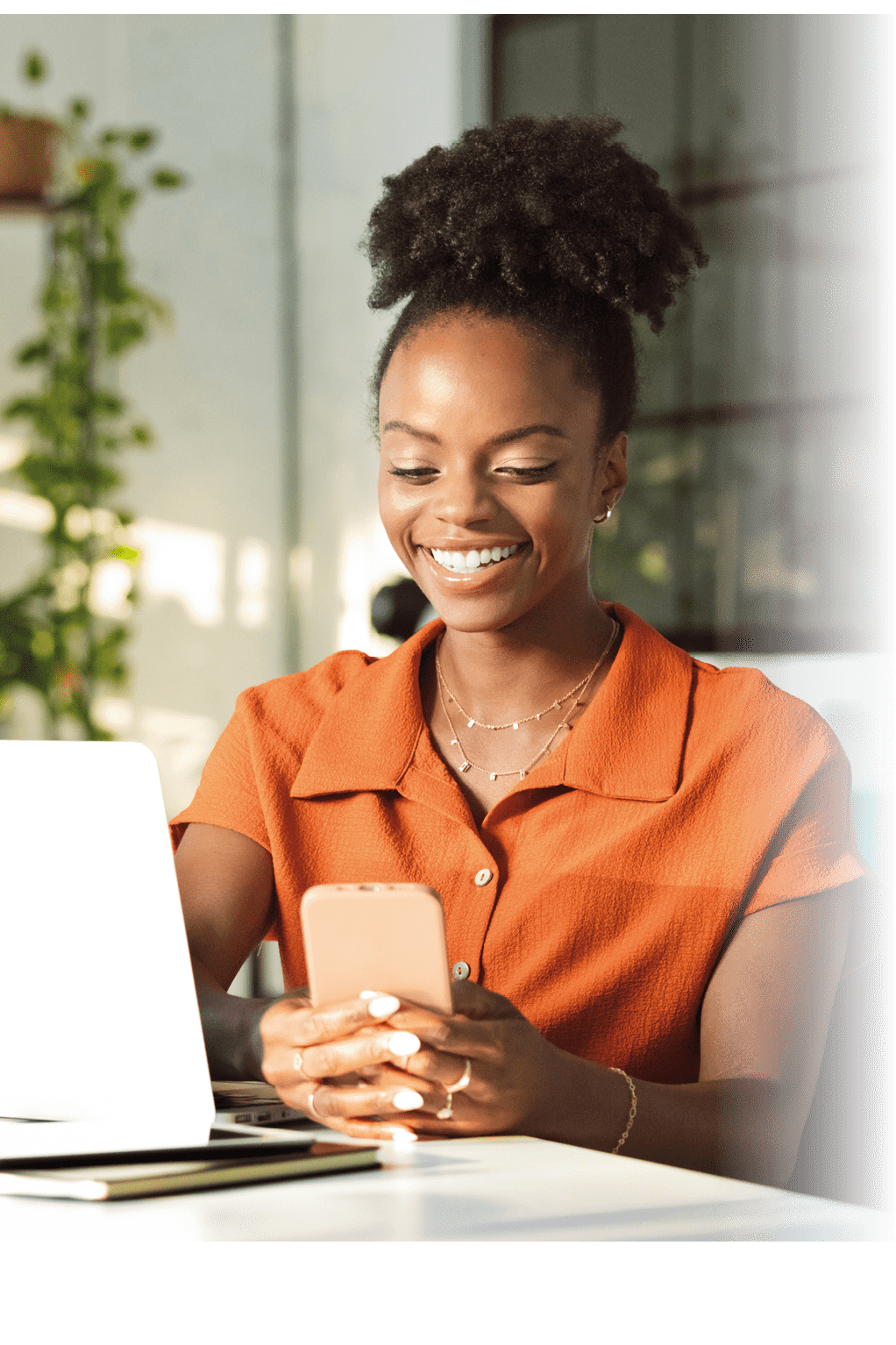 Beautiful, black women wearing orange dress sitting at the table in an office over laptop and using smart phone.