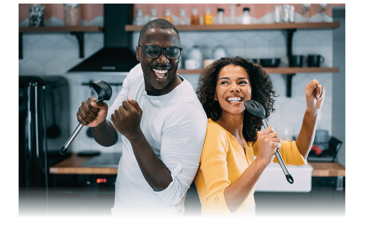 Shot of happy couple having fun during cooking in the kitchen at home. Lovely african-american couple holding kitchen utensil and singing during a cooking.