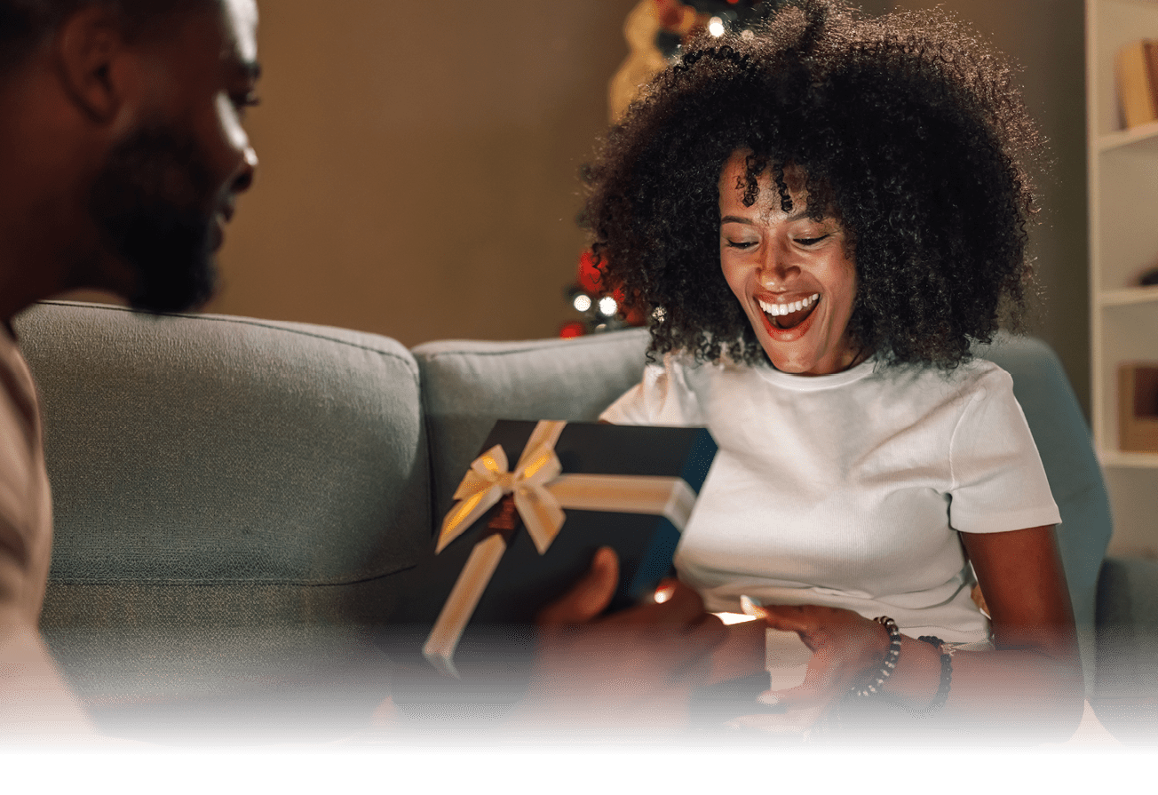 A delighted multiracial couple cherishes a tender gift exchange moment under a stunningly adorned Christmas tree, reflecting their warm connection and festive spirit in a comforting ambiance.
