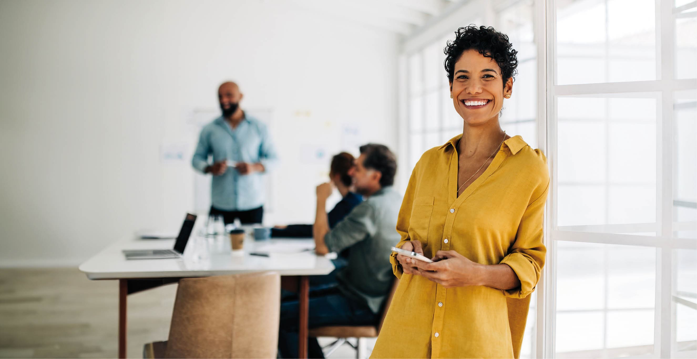 Female professional standing in an office and using a cellphone. Black business woman smiling at the camera in a boardroom. Colleagues having a meeting in the background.