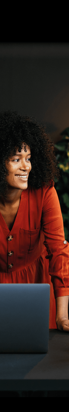 Cheerful businesswoman leaning over her desk and looking away while working on a laptop in a modern office