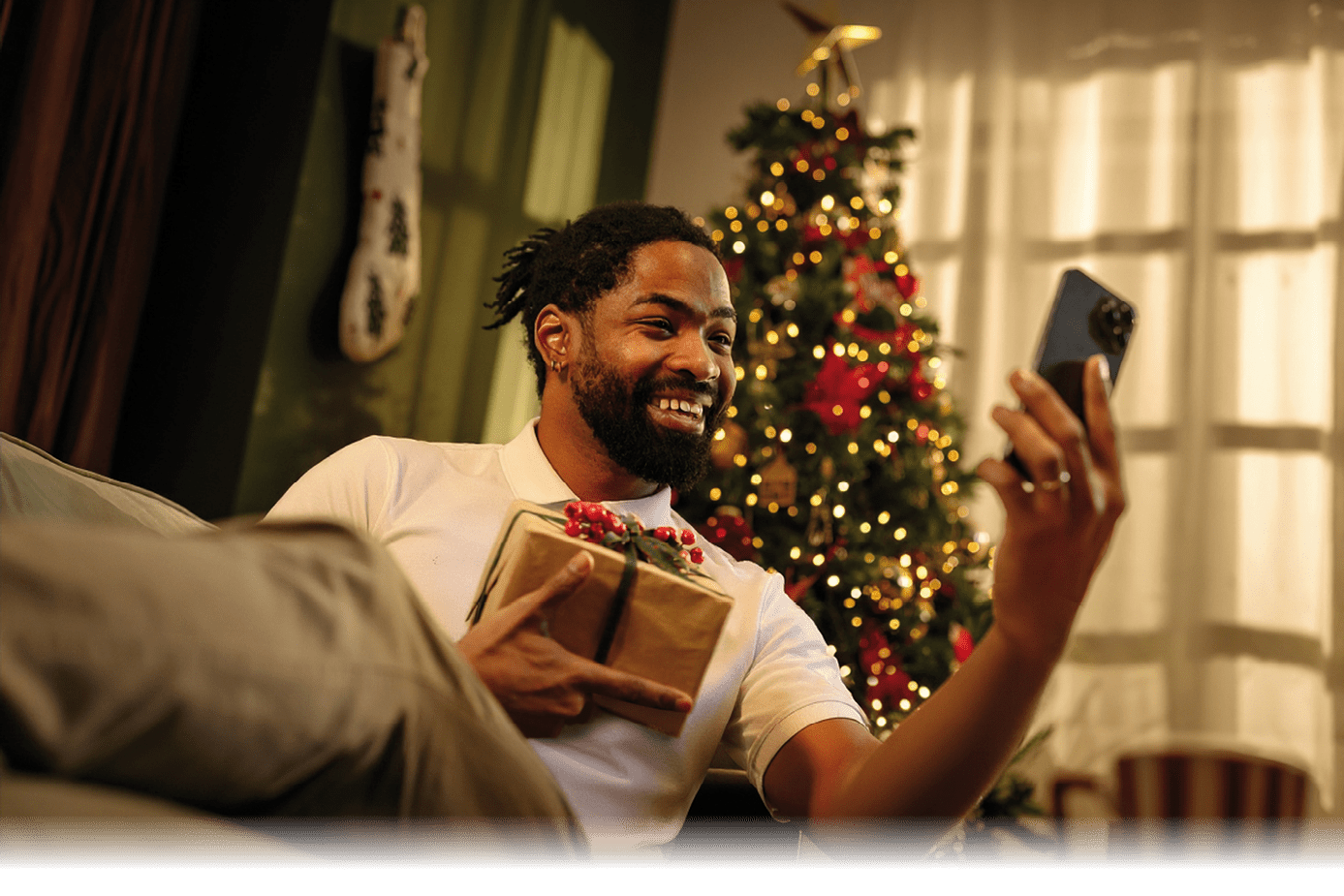Young man taking a selfie with a smartphone while holding a Christmas present, sitting on a sofa with a decorated tree behind