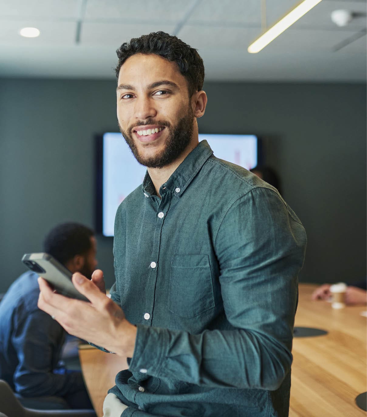 A confident businessman wearing a casual shirt leans on a conference table while holding his phone, with colleagues discussing work in the background.